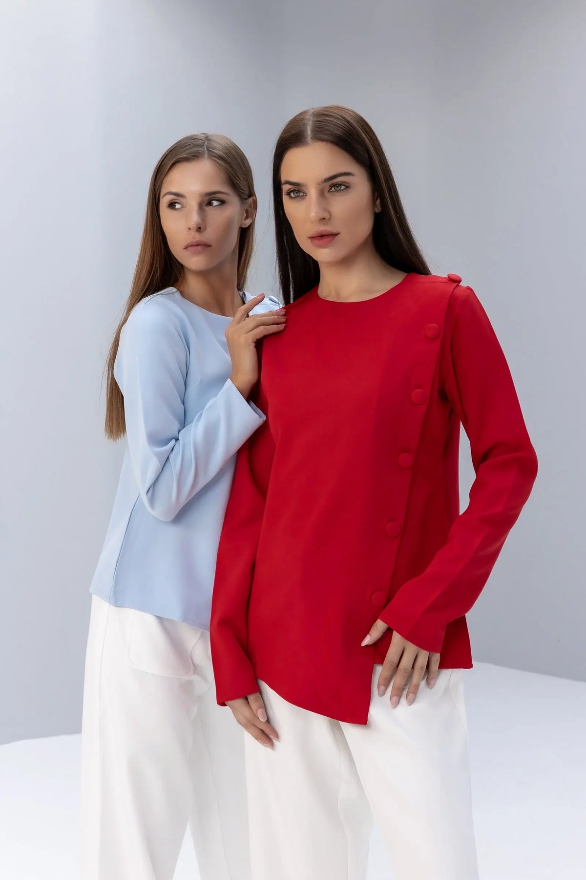 Two women standing side by side, one in a red top and white pants, the other in a light blue top and white pants, on a plain background.